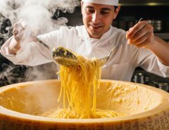 Chef tossing hot spaghetti inside a hollowed Parmigiano-Reggiano cheese wheel with steam rising