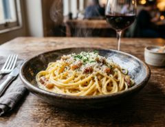 A steaming bowl of spaghetti carbonara garnished with black pepper and Parmesan on a rustic wooden table